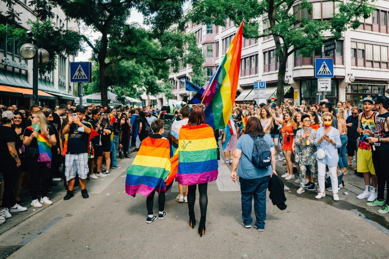 photo of 3 people's backs at a pride march, with rainbow flags as capes